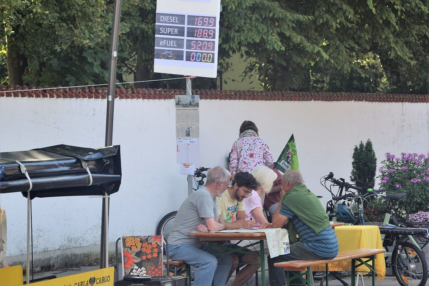 Personen auf Biertischgarnitur, davor eine Tankstellenpreistafel