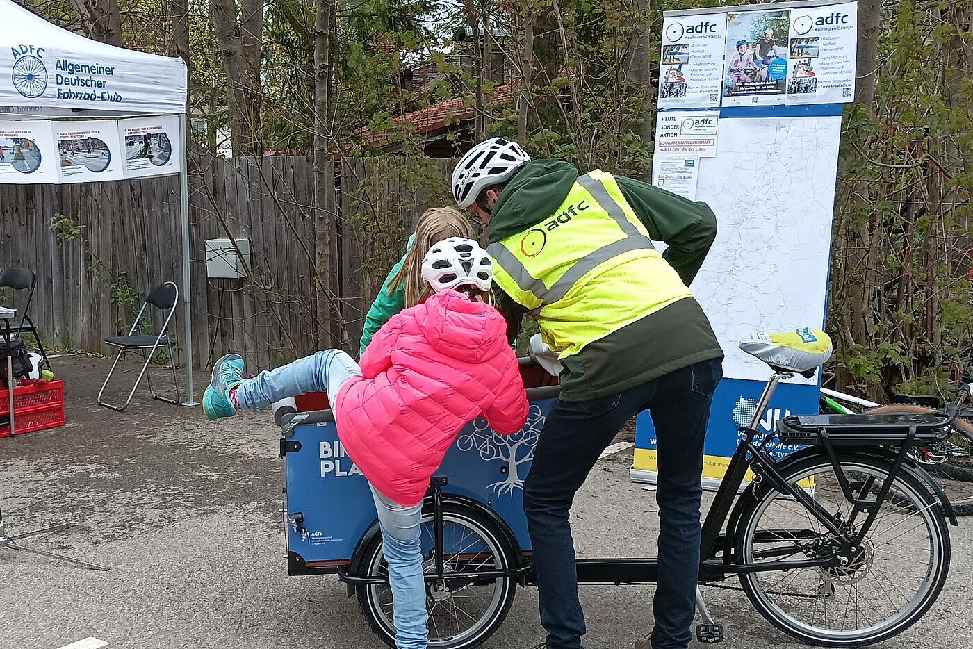 zwei Kinder steigen in ein Lastenrad, ein Mann hilft ihnen